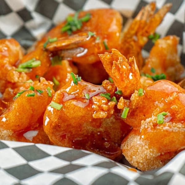 Crispy fried shrimp coated in a sweet and spicy orange sauce, garnished with fresh chives in a checkered basket.