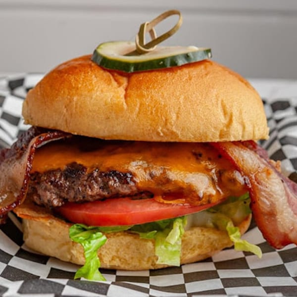 Mic's Kitchen bacon cheeseburger with lettuce, tomato, and a pickle spear on a checkered wrapper.