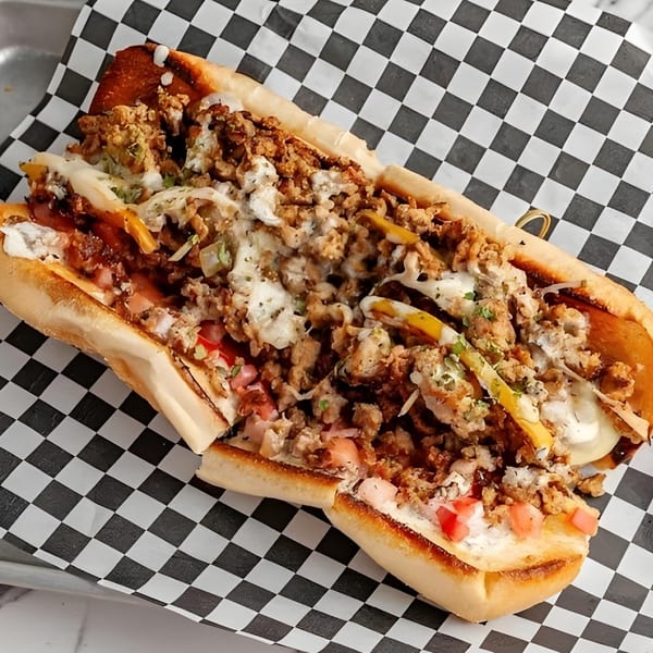 Mic's Kitchen's loaded hot dog with ground meat, cheese, tomatoes, and sauce on checkered paper.