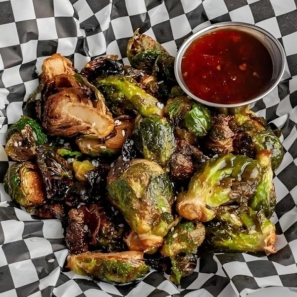 Crispy Brussels sprouts with sesame seeds and a side of sweet chili sauce in a checkered basket.