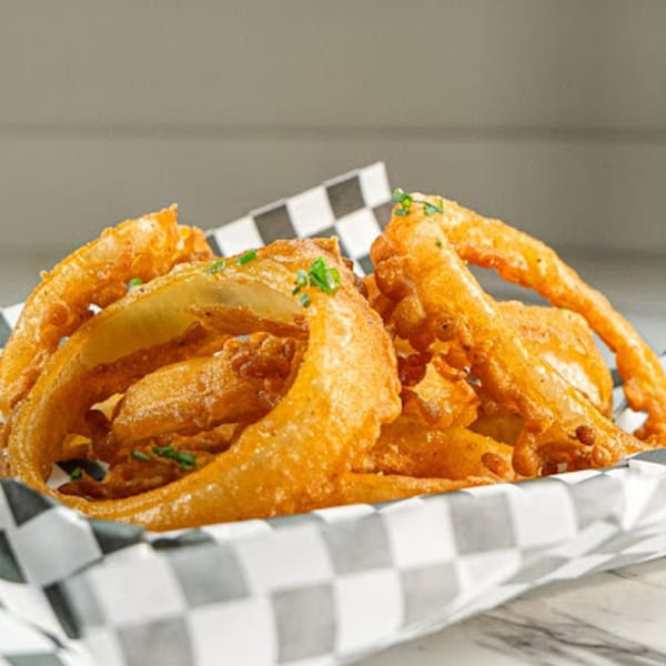 Crispy, golden-brown onion rings, lightly seasoned and garnished with fresh chives, served in a checkered paper basket.